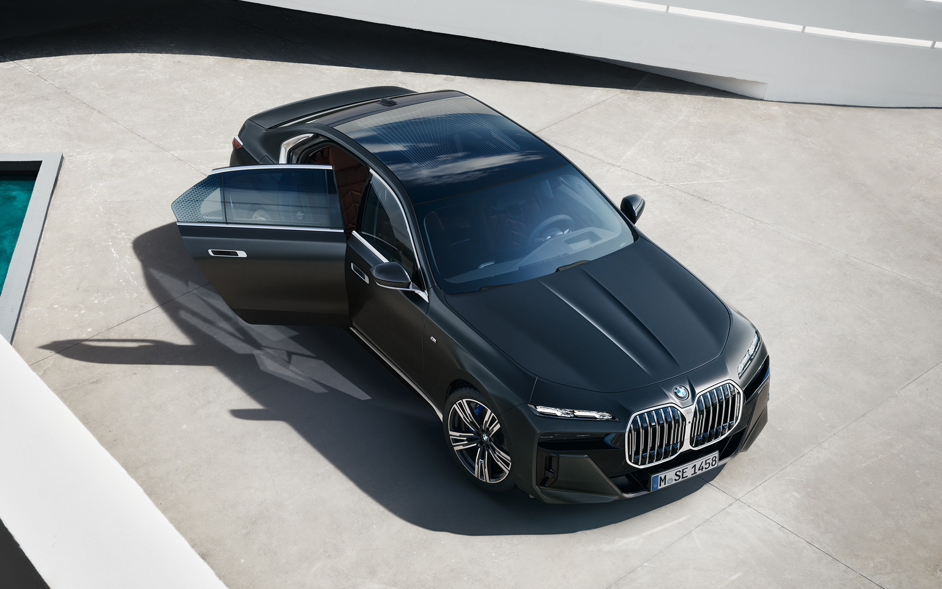 منظر علوي للسيارة BMW 7 Series Sedan G70 ICE 740i المجهزة بالباقة الرياضية M مع باب الراكب مفتوحاً، ويميزها لونها المعدني الرمادي القاتم فروزن